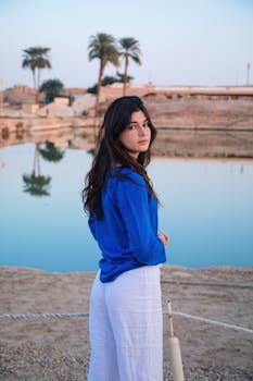 A woman in blue stands beside a tranquil pool reflecting palm trees in Luxor, Egypt.